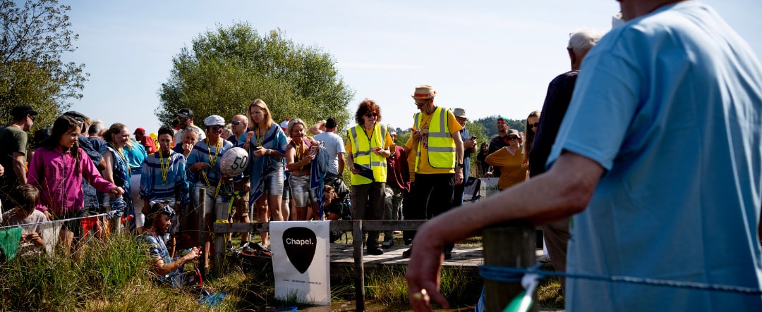 A sunlit crowd lined up along the bog trench beside a Chapel banner