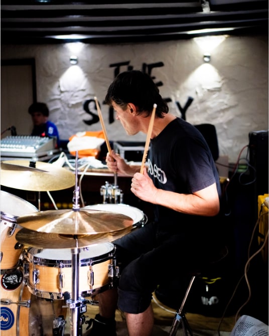 A drummer mid-song under spotlights at a village hall stage